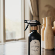 Black glass Mustard Seed multipurpose Gratitude Bottle with spray pump on a table near the window.