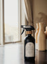 Black glass Mustard Seed multipurpose Gratitude Bottle with spray pump on a table near the window.