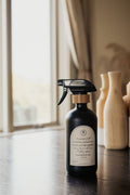 Black glass Mustard Seed multipurpose Gratitude Bottle with spray pump on a table near the window.