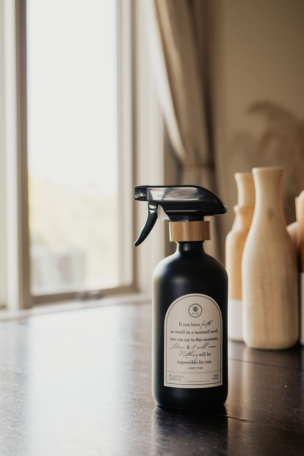Black glass Mustard Seed multipurpose Gratitude Bottle with spray pump on a table near the window.