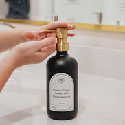 Black glass I Can Do All Things multipurpose Gratitude Bottle with gold pump being used by a bathroom sink.