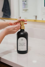 Black glass I Can Do All Things multipurpose Gratitude Bottle with gold pump being used by a bathroom sink.