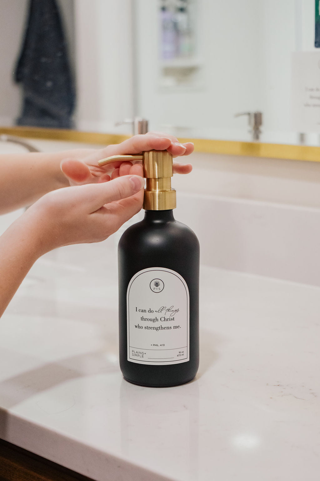 Black glass I Can Do All Things multipurpose Gratitude Bottle with gold pump being used by a bathroom sink.
