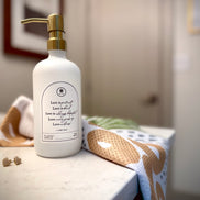 A gold pump on a white 16 oz glass reusable soap or lotion bottle with a printed label reading 'Love is patient, Love is kind, Love is always hopeful. Love never gives up, Love endures! 1 Cor. 13:4-7' next to a beautifully printed hand towel based on the Matthew 17 mustard seed verse.
