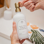 White glass I Can Do All Things multipurpose Gratitude Bottle with gold pump being used by the bathroom sink.