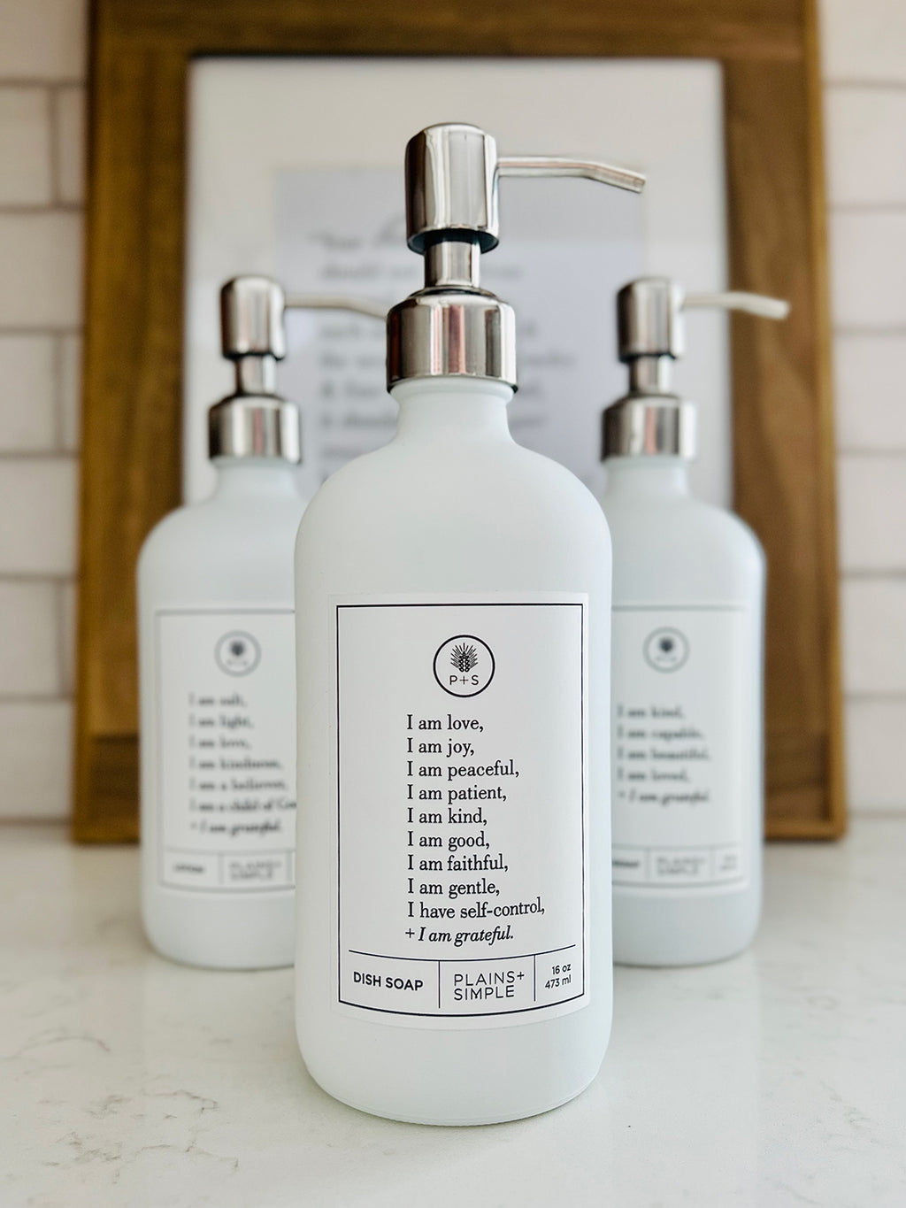 White glass Fruits of the Spirit dish soap Gratitude Bottle with silver pump sitting with the matching hand soap and lotion bottles on a kitchen counter.