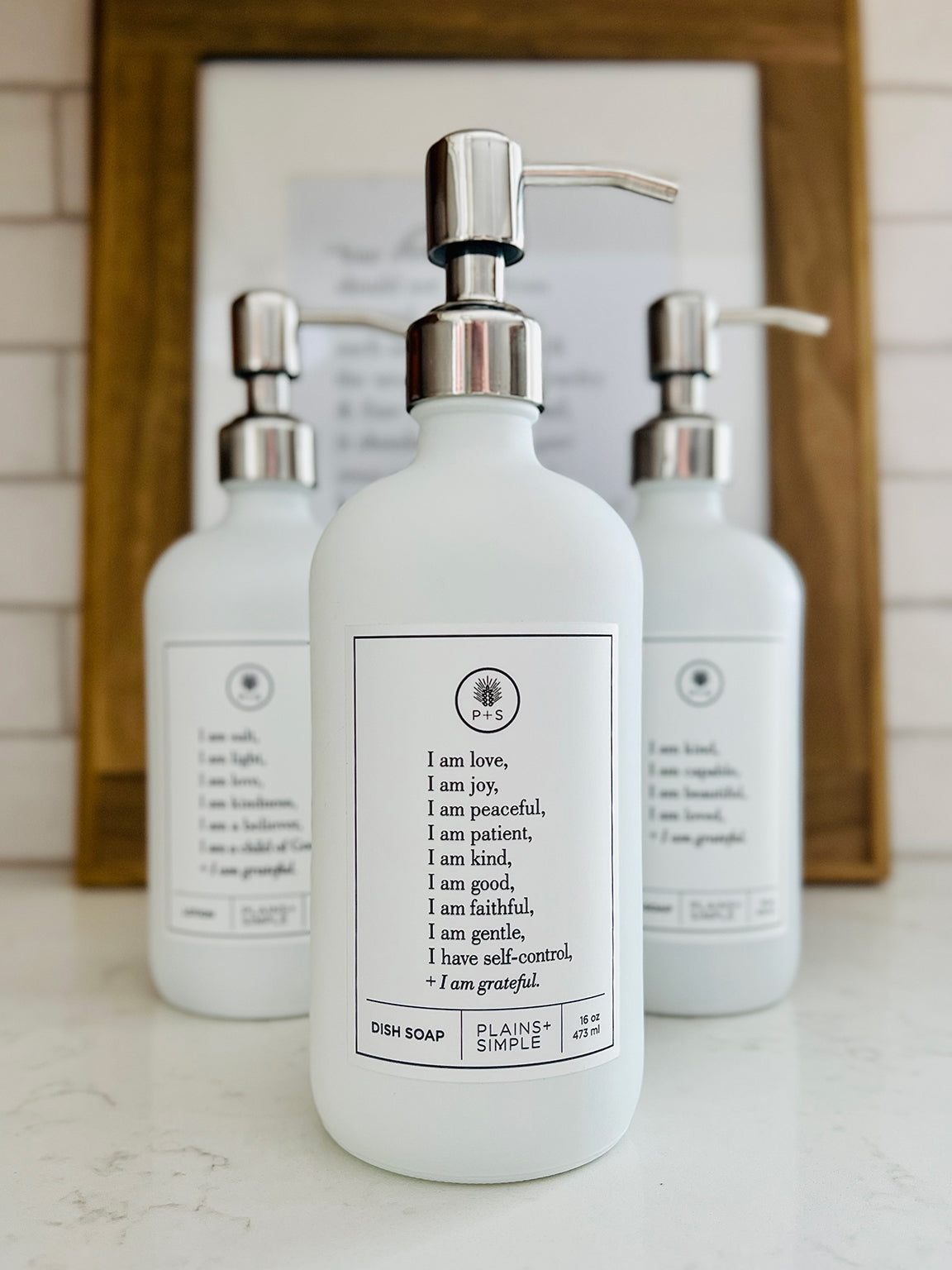 White glass Fruits of the Spirit dish soap Gratitude Bottle with silver pump sitting with the matching hand soap and lotion bottles on a kitchen counter.