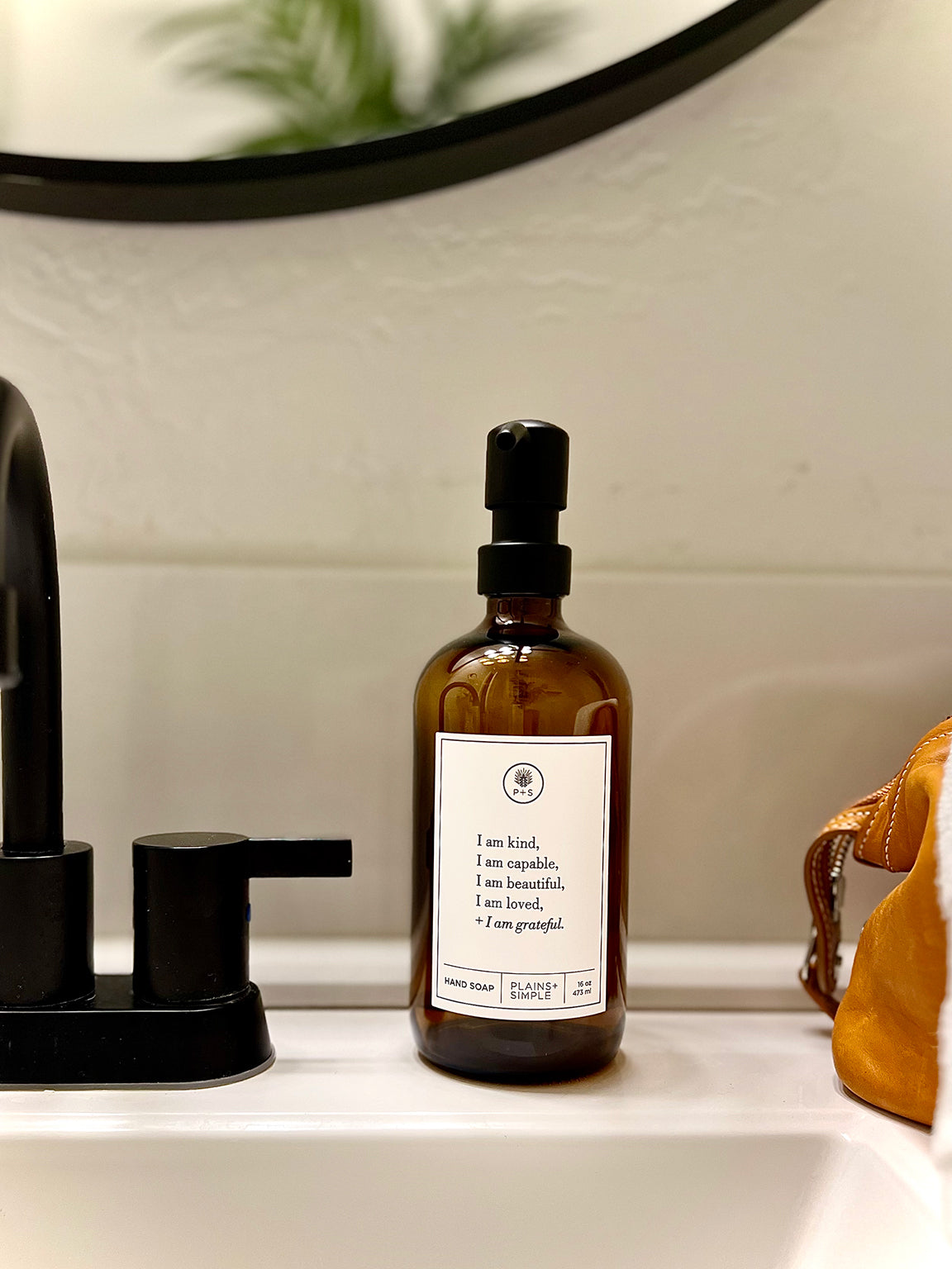 Amber glass I am Kind hand soap  Gratitude Bottle with black pump next to bathroom sink.