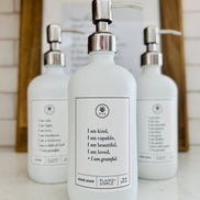 White glass I am Kind hand soap Gratitude Bottle with silver pump with matching dish soap and lotion bottles on the kitchen counter.