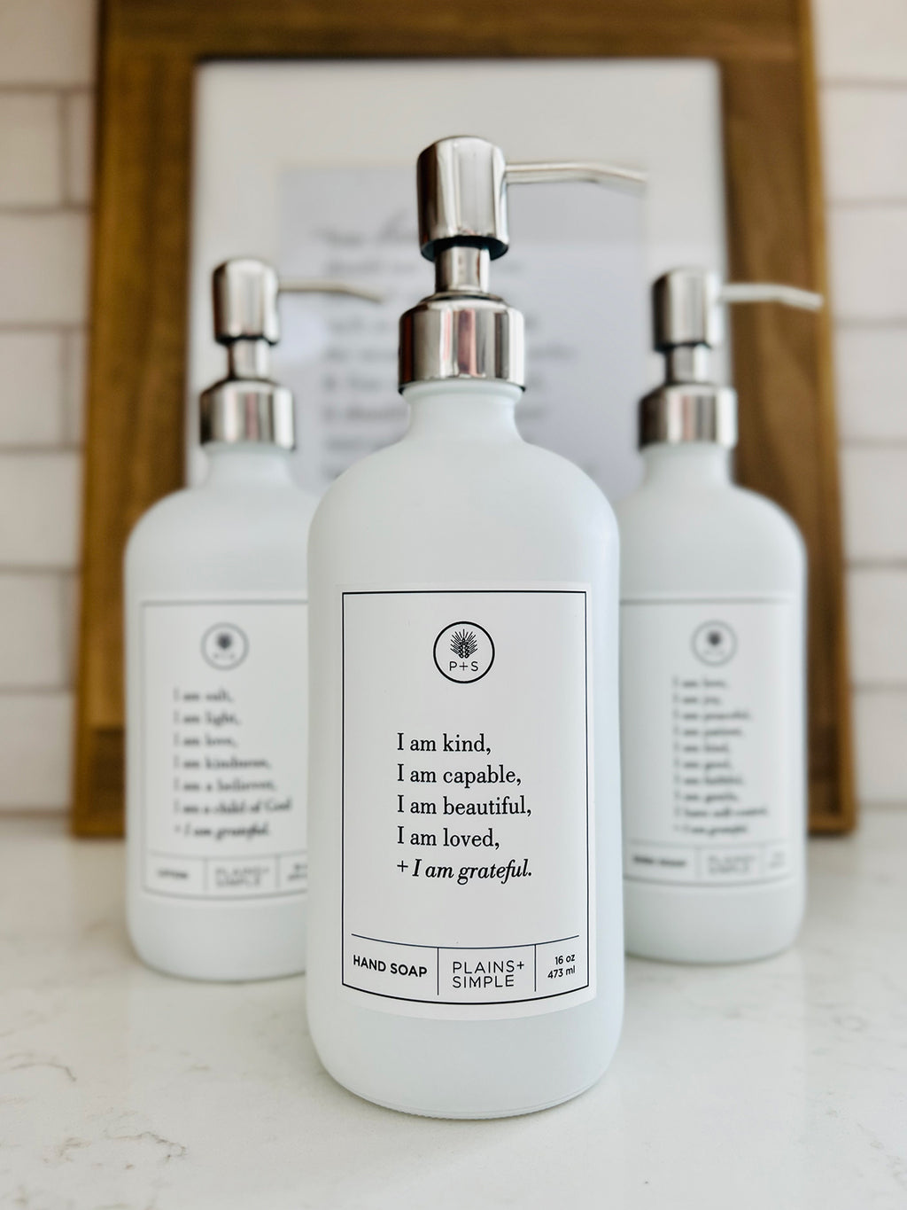 White glass I am Kind hand soap Gratitude Bottle with silver pump with matching dish soap and lotion bottles on the kitchen counter.