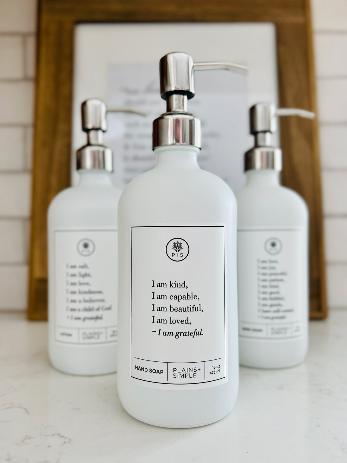 White glass I am Kind hand soap Gratitude Bottle with silver pump with matching dish soap and lotion bottles on the kitchen counter.