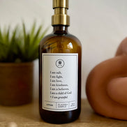 Amber glass I am Salt & Light lotion Gratitude Bottle with gold pump on a bathroom shelf with jewelry.