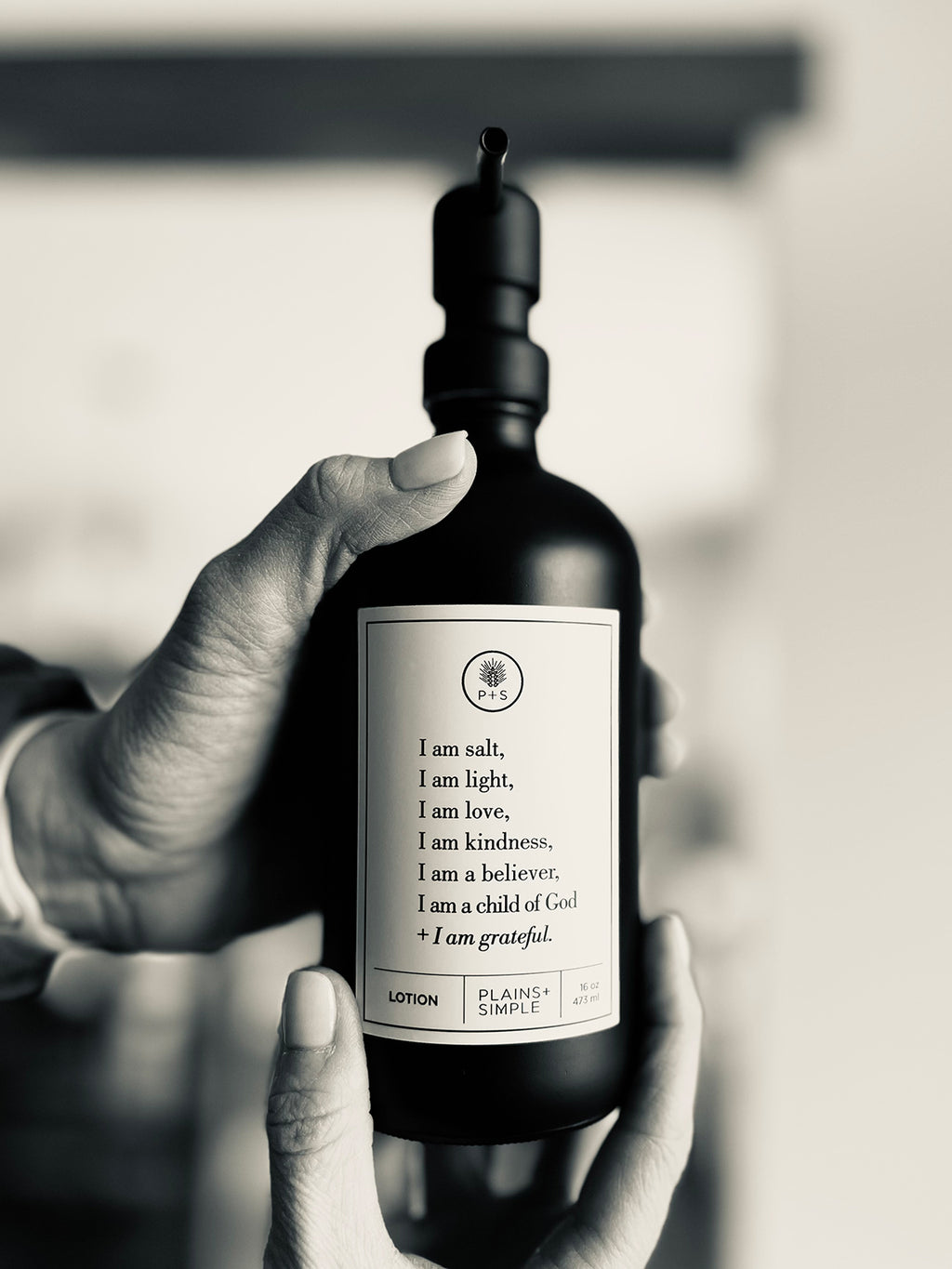 Black glass I am Salt & Light lotion Gratitude Bottle with black pump being held by hands.