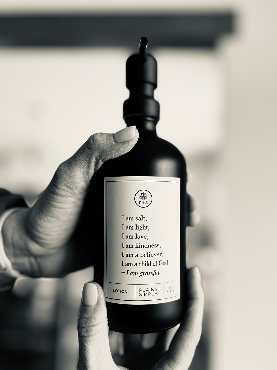 Black glass I am Salt & Light lotion Gratitude Bottle with black pump being held by hands.