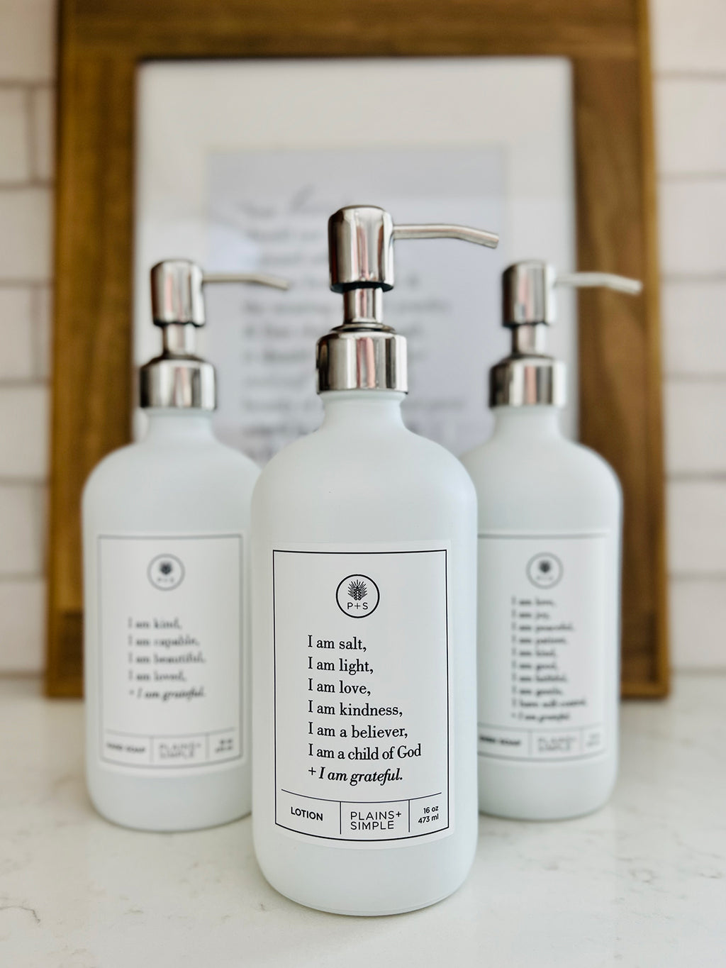 White glass I am Salt & Light lotion Gratitude Bottle with silver pump shown with matching hand soap and dish soap bottles on the kitchen counter.