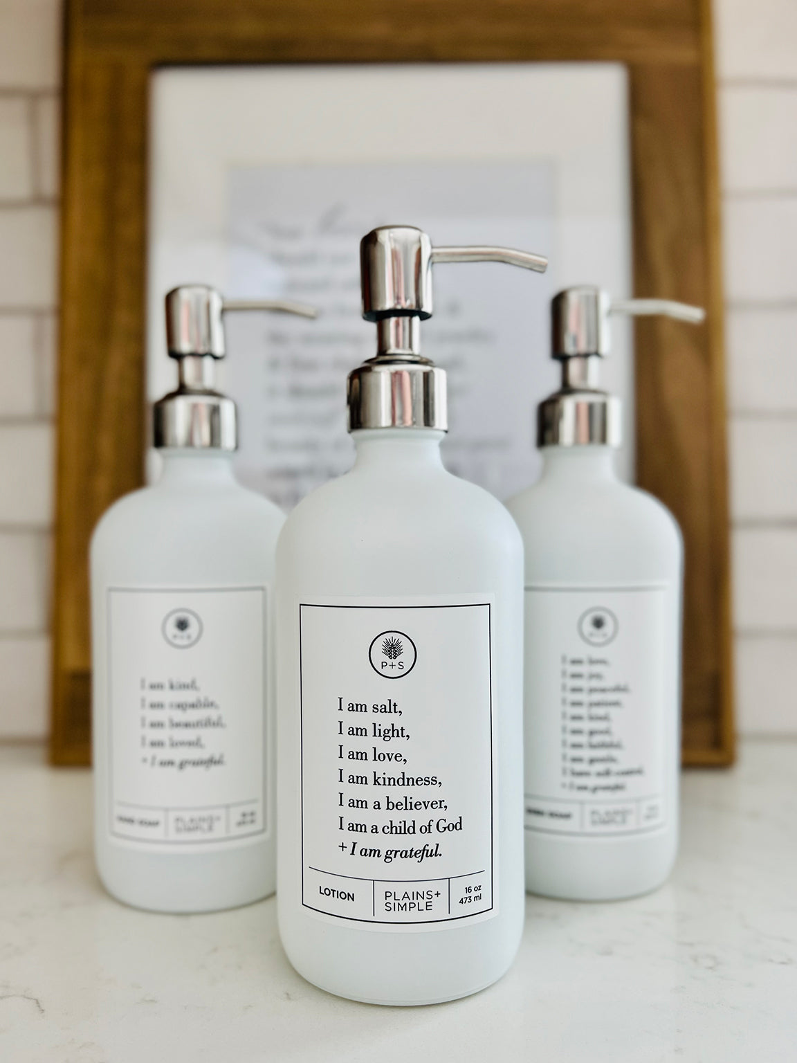 White glass I am Salt & Light lotion Gratitude Bottle with silver pump shown with matching hand soap and dish soap bottles on the kitchen counter.