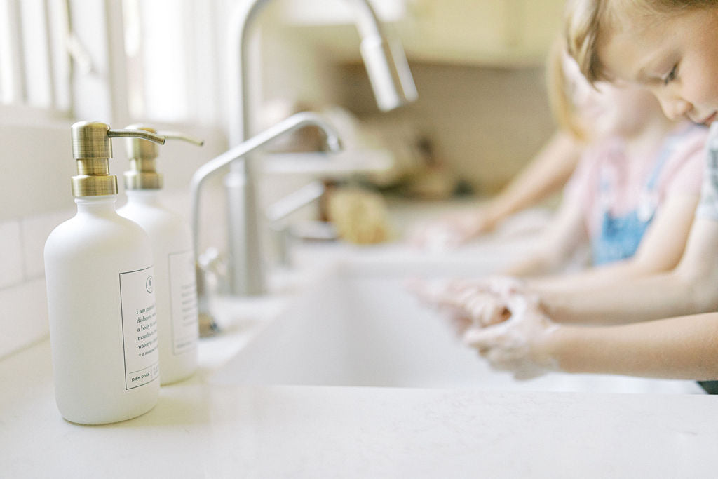 Children washing hands with soap while pausing with gratitude at a sink, with two reusable gratitude soap and lotion bottles in the foreground.
