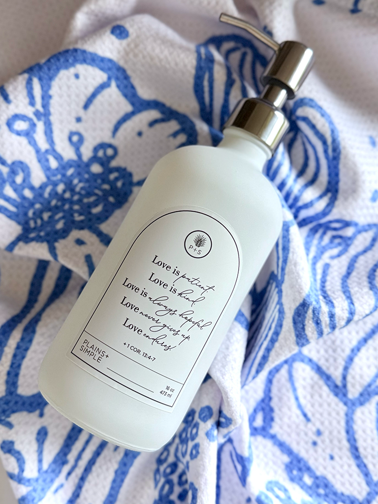 White glass Love Is Gratitude Bottle with silver pump on a blue and white floral patterned hand towel.