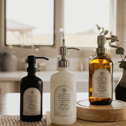 Three Trust multipurpose glass Gratitude Bottles with pump dispensers on a wooden surface in a kitchen setting.