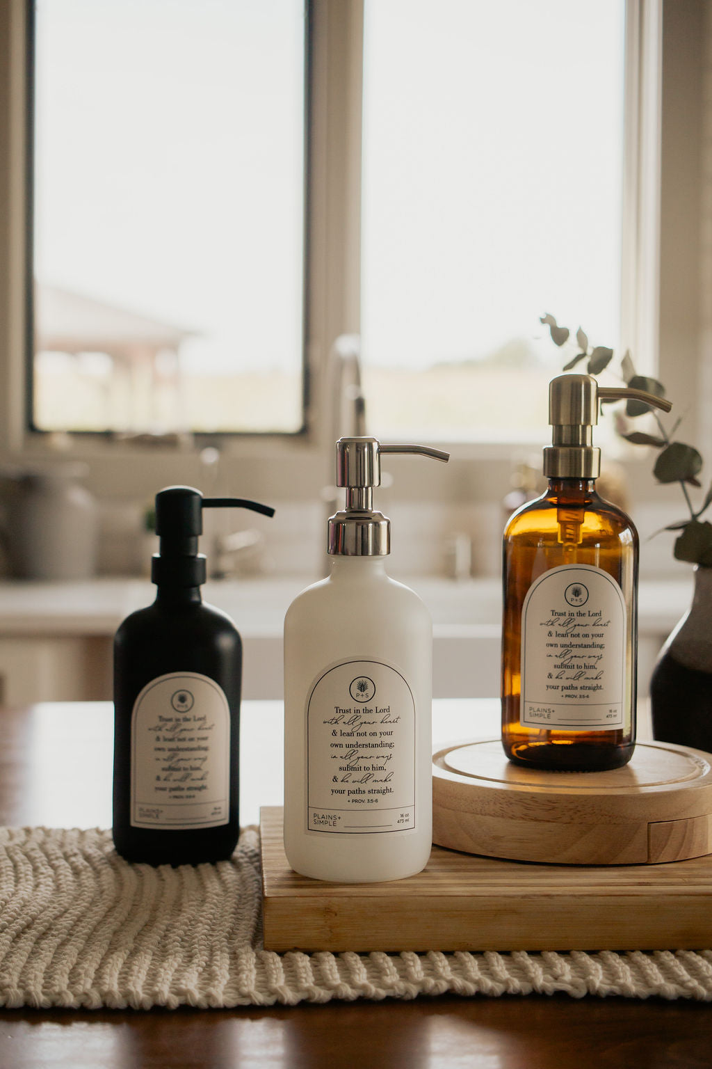 Three Trust multipurpose glass Gratitude Bottles with pump dispensers on a wooden surface in a kitchen setting.